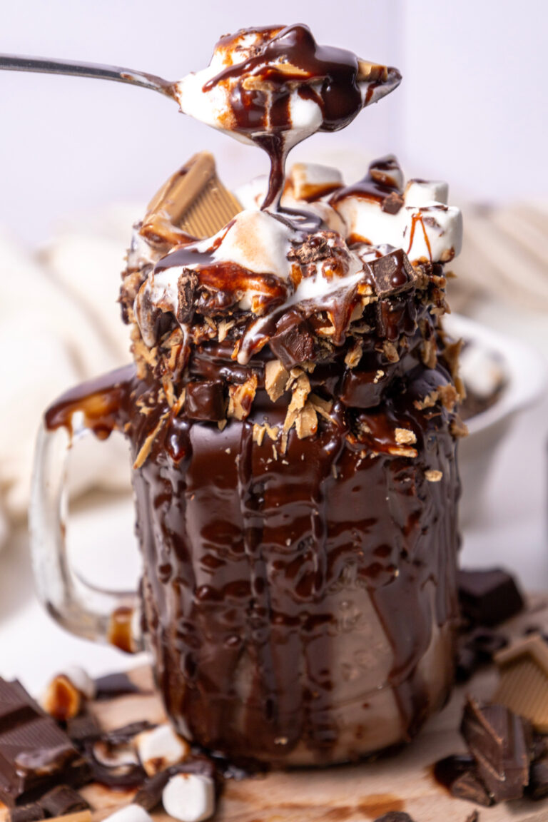 A mason jar filled with a chocolate milkshake, topped with marshmallows, chocolate sauce, and chocolate pieces, with a spoon lifting a marshmallow from the top.