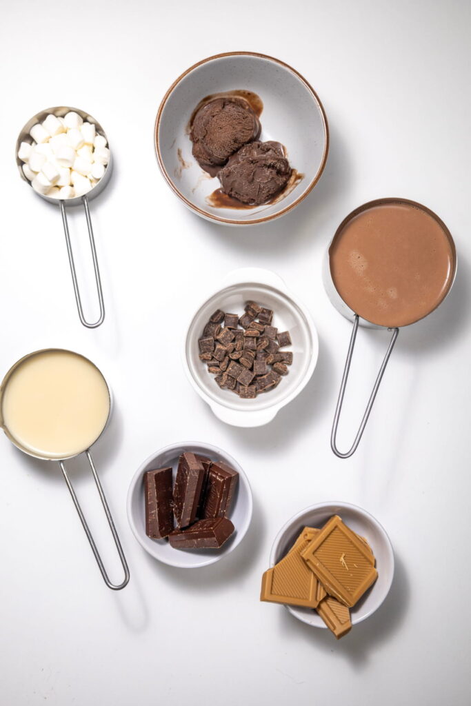 Various dessert ingredients in bowls and measuring cups arranged on a white surface, including marshmallows, chocolate, ice cream, and condensed milk.