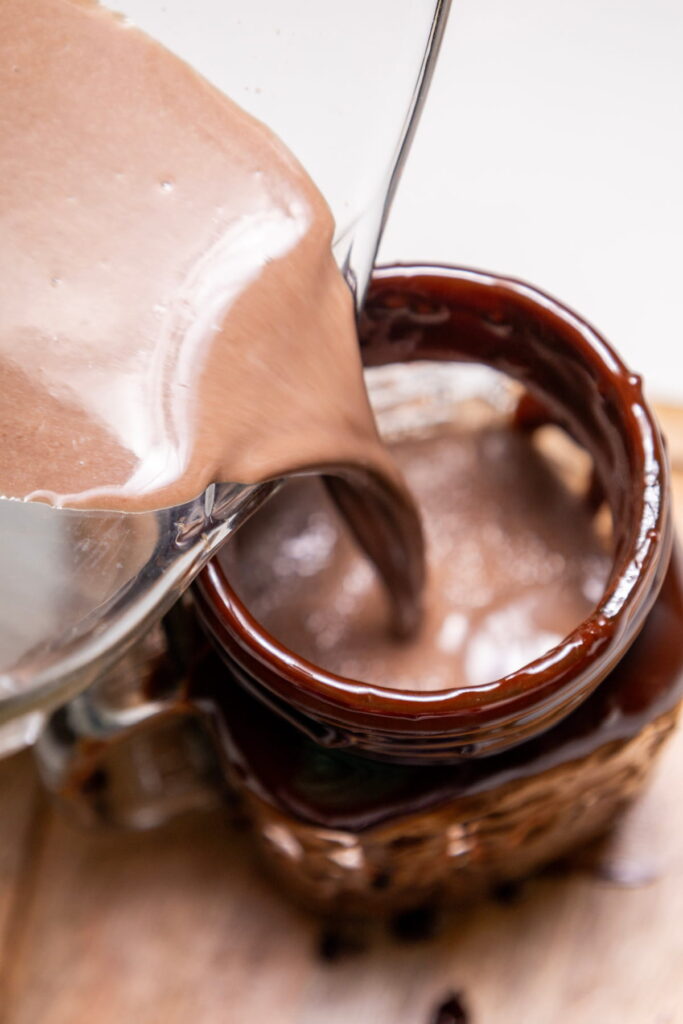 A thick chocolate beverage is being poured from a glass container into a brown mug on a wooden surface.