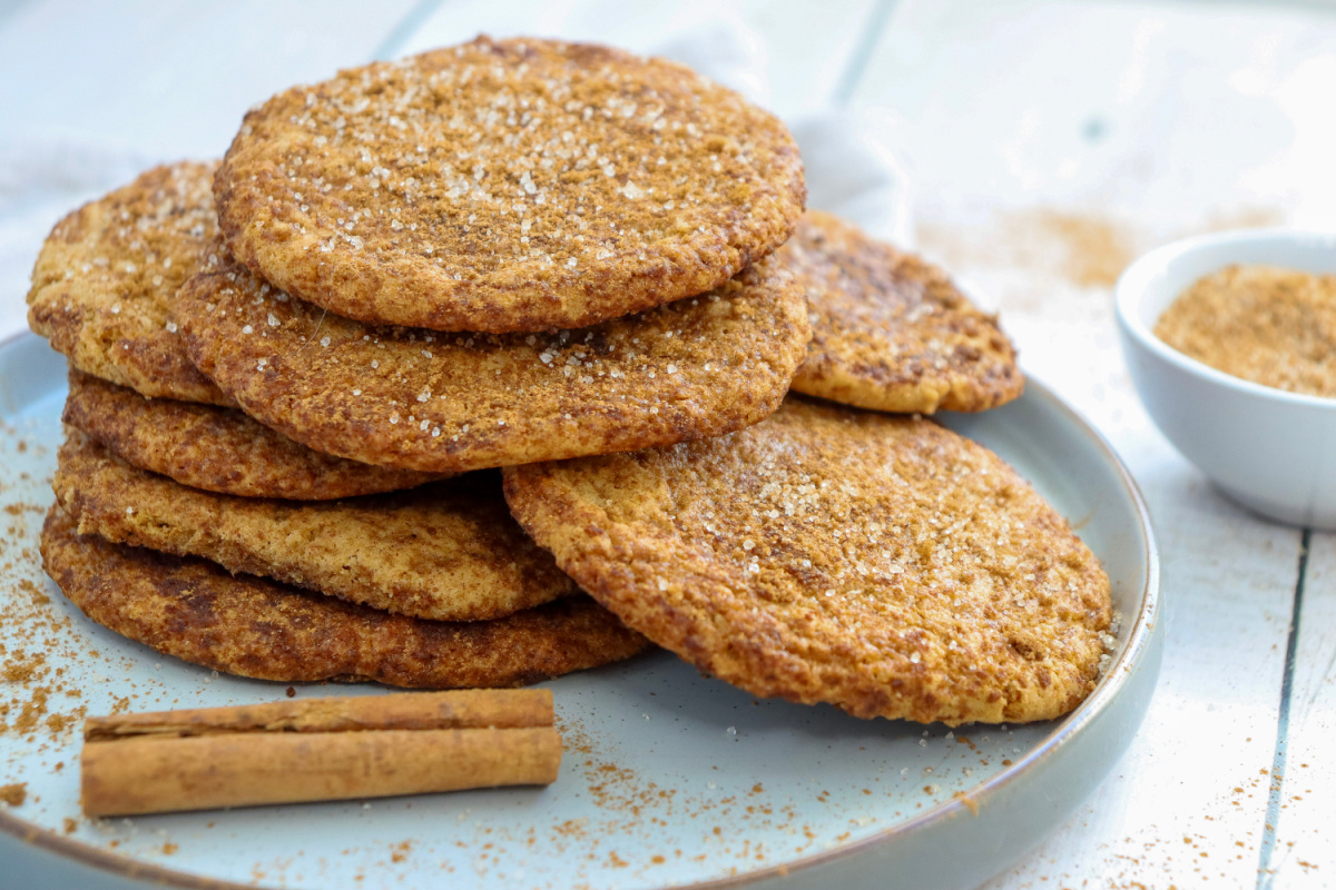 A stack of snickerdoodle cookies sprinkled with sugar on a plate, with a cinnamon stick and a bowl of cinnamon sugar nearby.