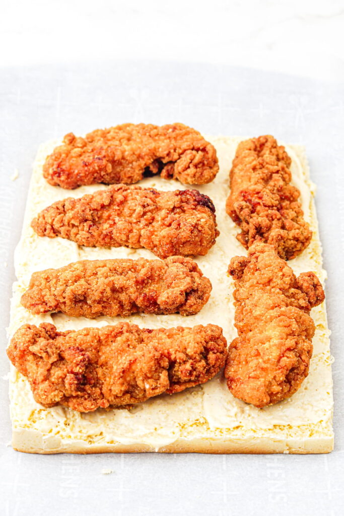 Six breaded chicken tenders are arranged in two rows on a rectangular slice of bread spread with a creamy sauce.