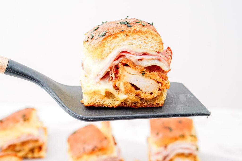 A spatula holds a slider sandwich filled with fried chicken, cheese, and deli meat on a white background.