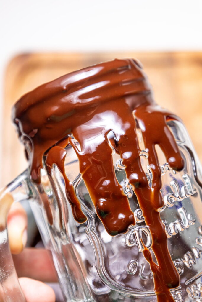 A clear glass jar is tilted as thick chocolate sauce drips down the rim and sides.