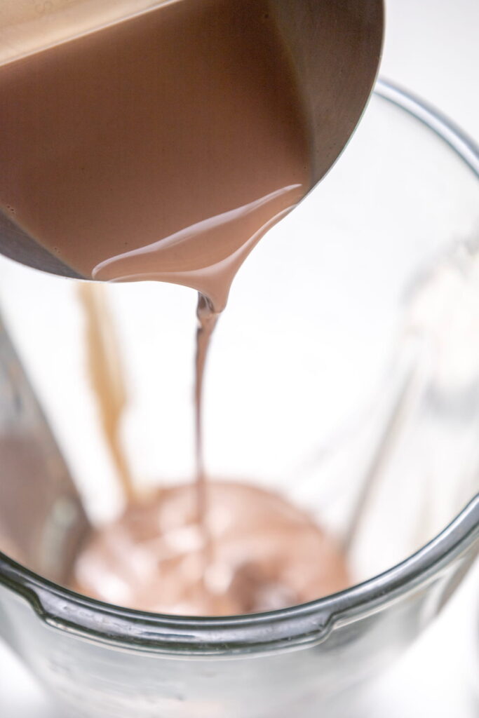 Chocolate liquid being poured from a metal container into a glass blender jar.
