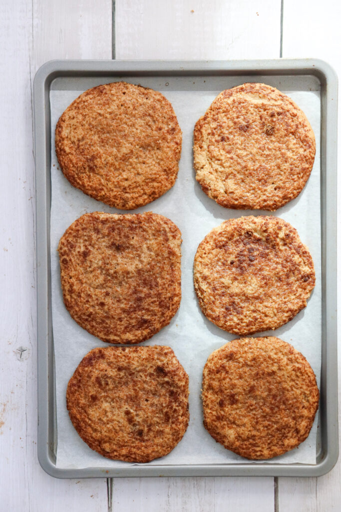 A baking tray lined with parchment paper holds six large, round, baked flatbread pieces arranged in two columns of three.
