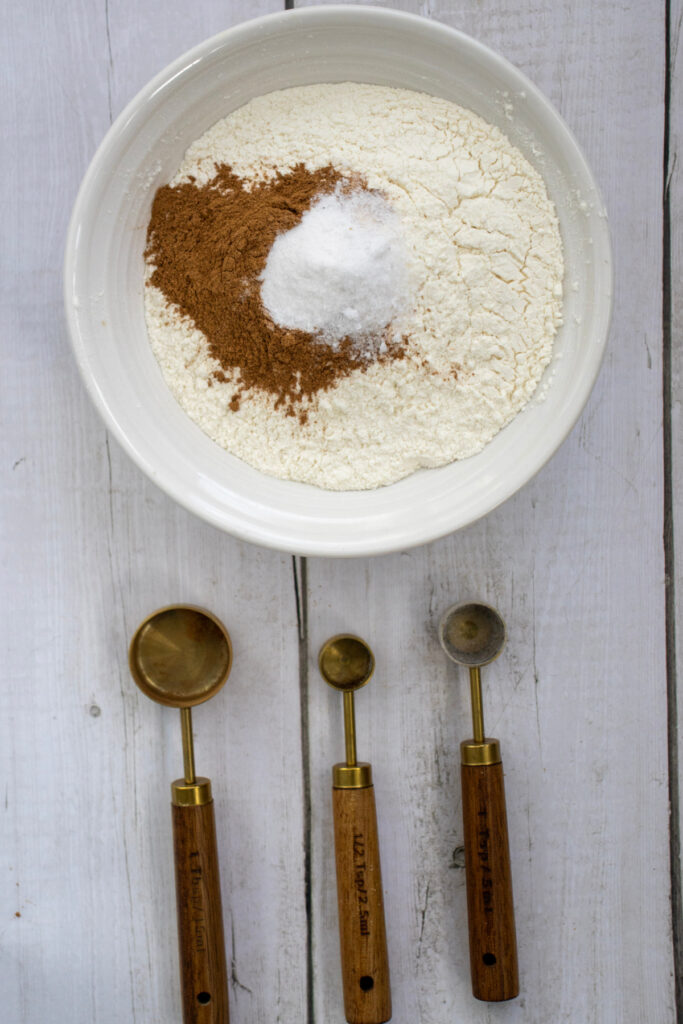 A white bowl with flour, baking soda, and cinnamon sits on a wooden surface, with three measuring spoons placed below it.