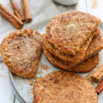 A plate of cinnamon-dusted flatbreads sits on a white table with a bowl of cinnamon sugar and whole cinnamon sticks nearby.
