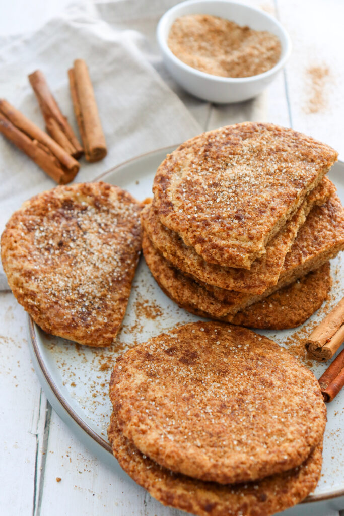 A plate of cinnamon-dusted flatbreads sits on a white table with a bowl of cinnamon sugar and whole cinnamon sticks nearby.