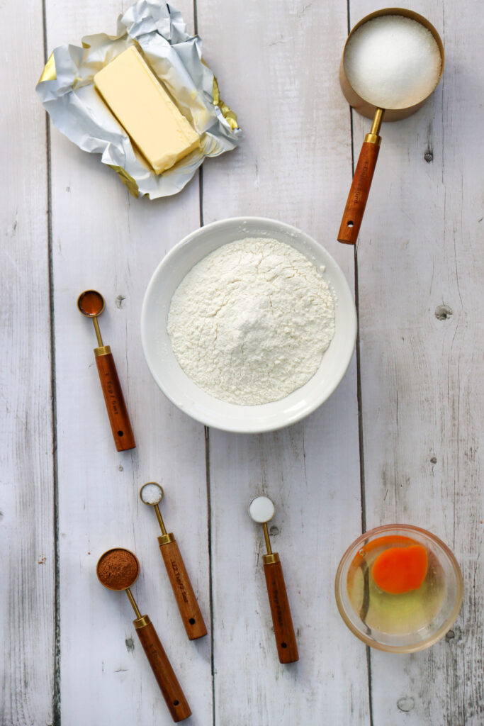 Baking ingredients on a wooden surface: butter, sugar, flour in a bowl, an egg in a glass bowl, cocoa powder, and measuring spoons.