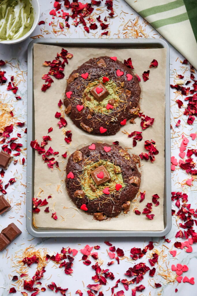 Two decorated cookies with green frosting, square chocolates, and red heart sprinkles on a parchment-lined baking tray, surrounded by dried rose petals and chocolate pieces.
