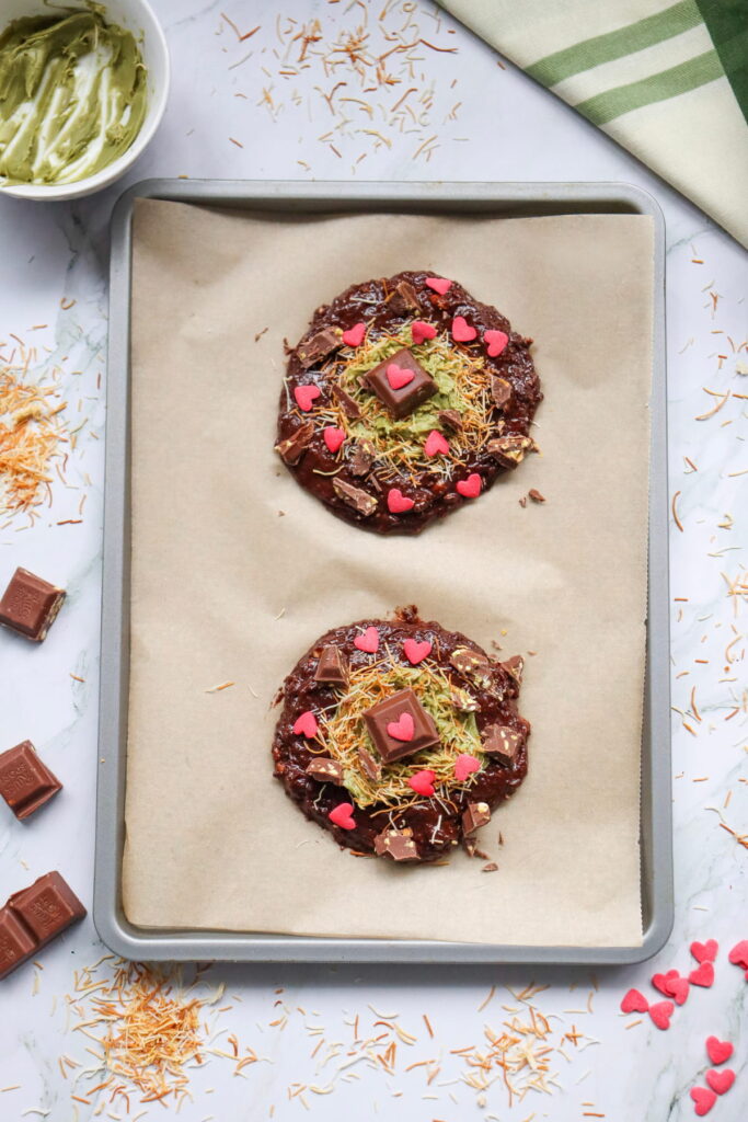 Two chocolate cookies topped with shredded coconut, heart-shaped sprinkles, and chocolate pieces on a parchment-lined baking tray. Baking ingredients and a green towel are nearby.