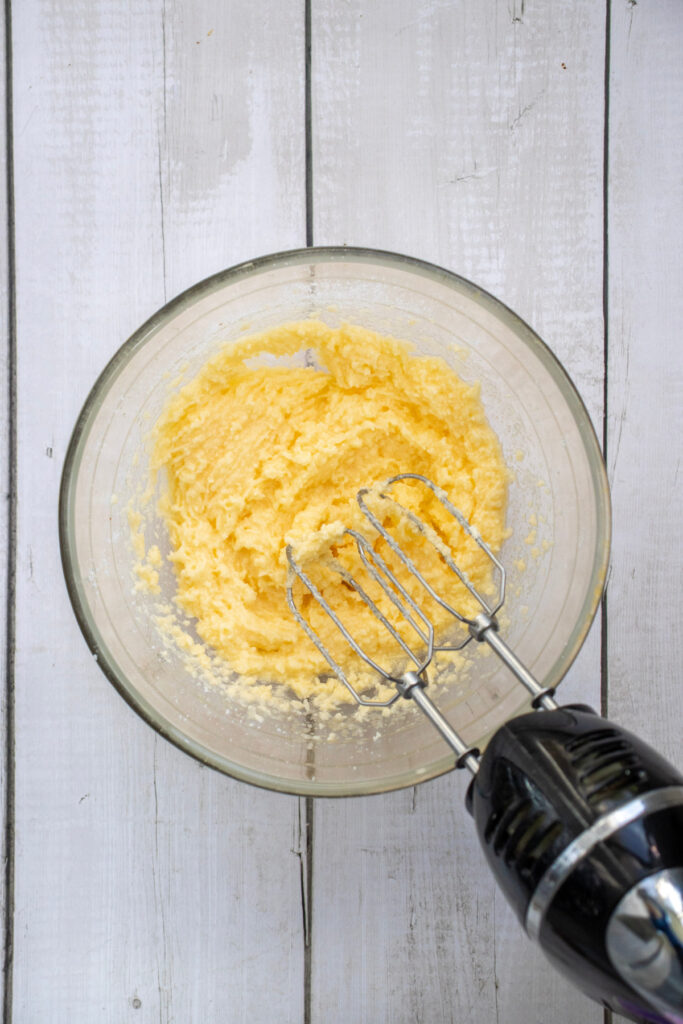A glass bowl with creamed butter and sugar, partially mixed, sits on a white wooden surface with a handheld electric mixer inside.