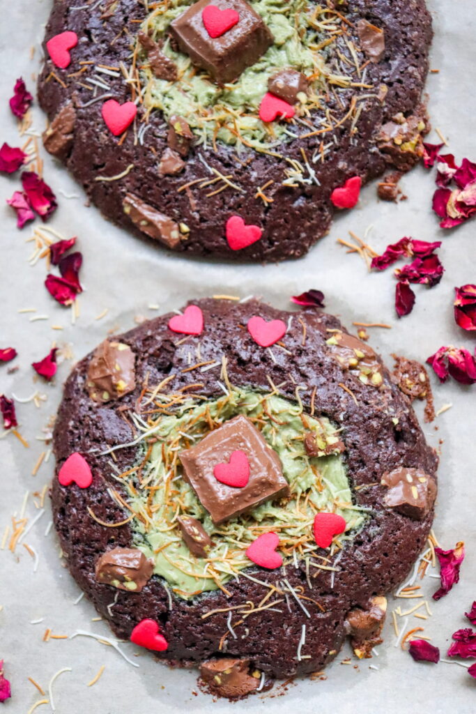 Two large chocolate cookies with green frosting centers, topped with chocolate pieces, red heart sprinkles, shredded coconut, and surrounded by dried rose petals on parchment paper.