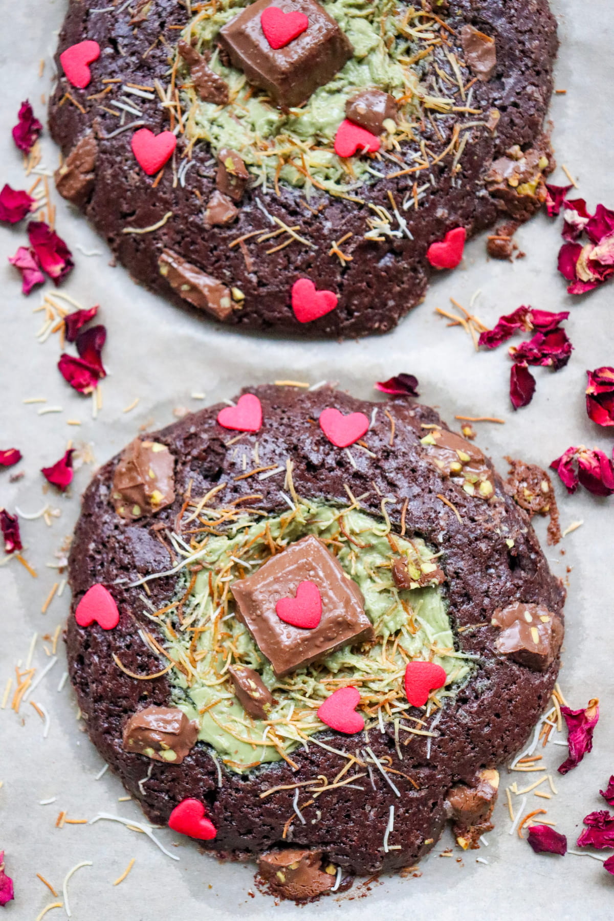 Two large chocolate cookies with green frosting centers, topped with chocolate pieces, red heart sprinkles, shredded coconut, and surrounded by dried rose petals on parchment paper.