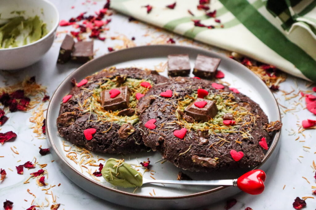 Two large chocolate cookies topped with red heart decorations and chocolate pieces on a plate, surrounded by rose petals and a small bowl of green spread.