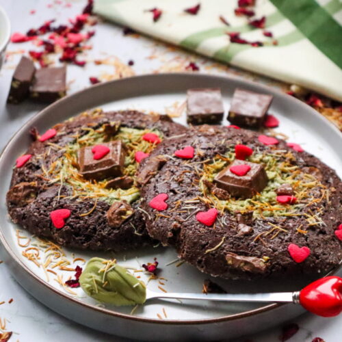 Two large chocolate cookies topped with red heart decorations and chocolate pieces on a plate, surrounded by rose petals and a small bowl of green spread.
