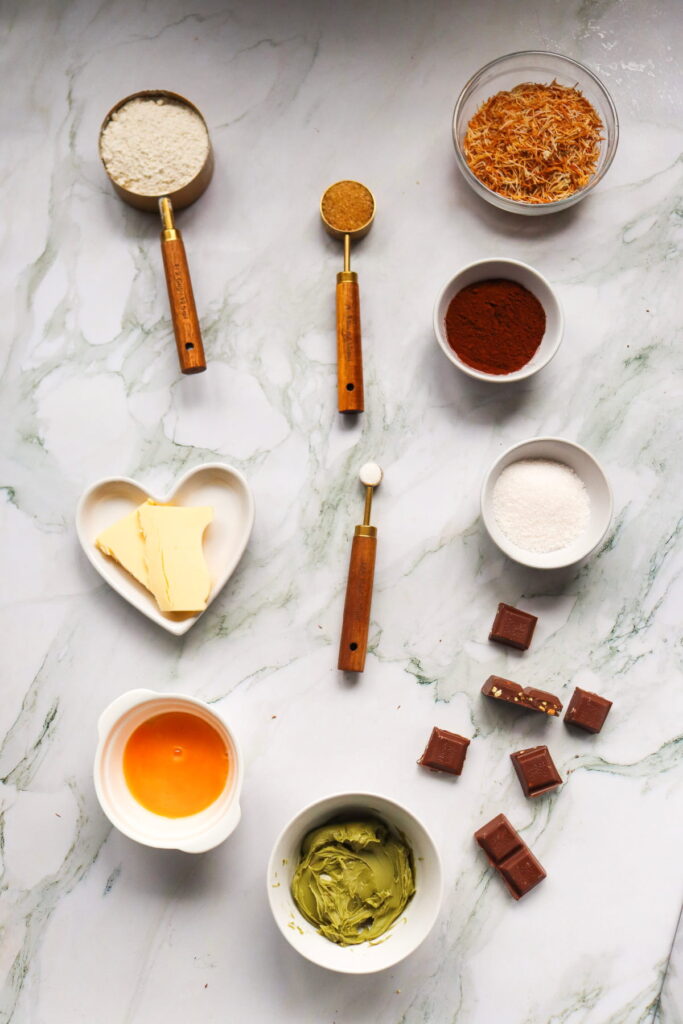 Various baking ingredients—flour, sugar, butter, egg, matcha paste, cocoa powder, brown sugar, shredded coconut, and chocolate pieces—are arranged on a marble countertop.