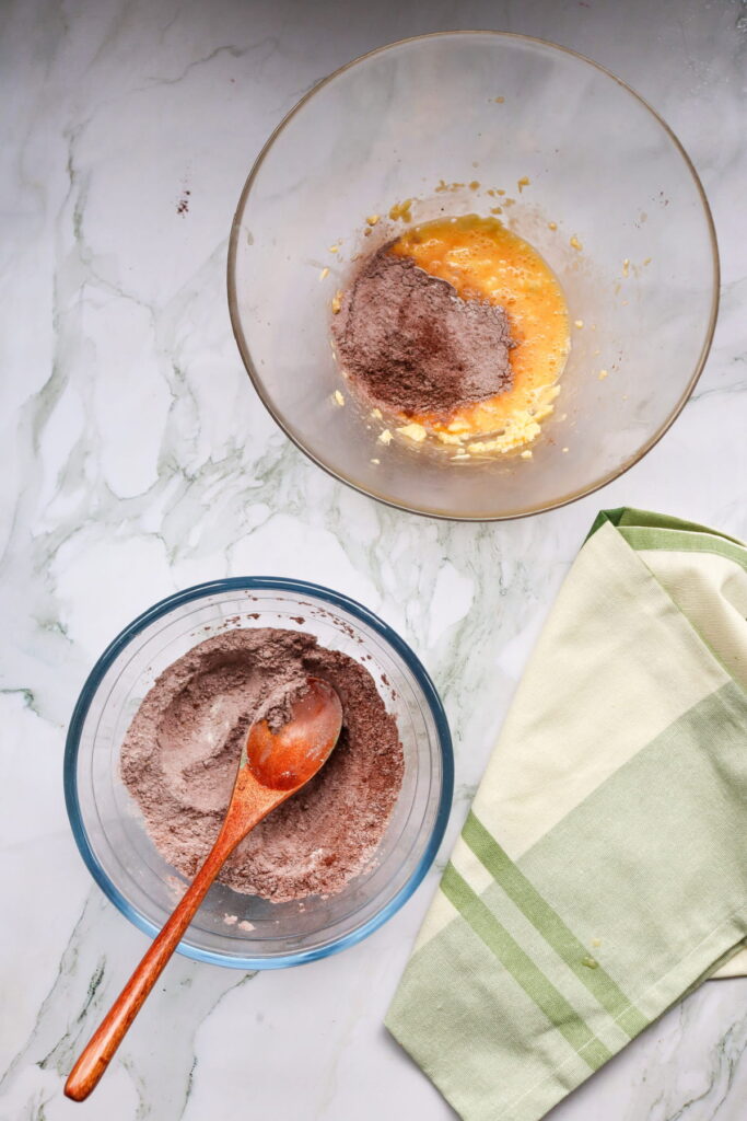 Two glass bowls on a marble surface; one with a chocolate powder mixture and wooden spoon, the other with wet ingredients and cocoa powder. A green striped kitchen towel is beside them.