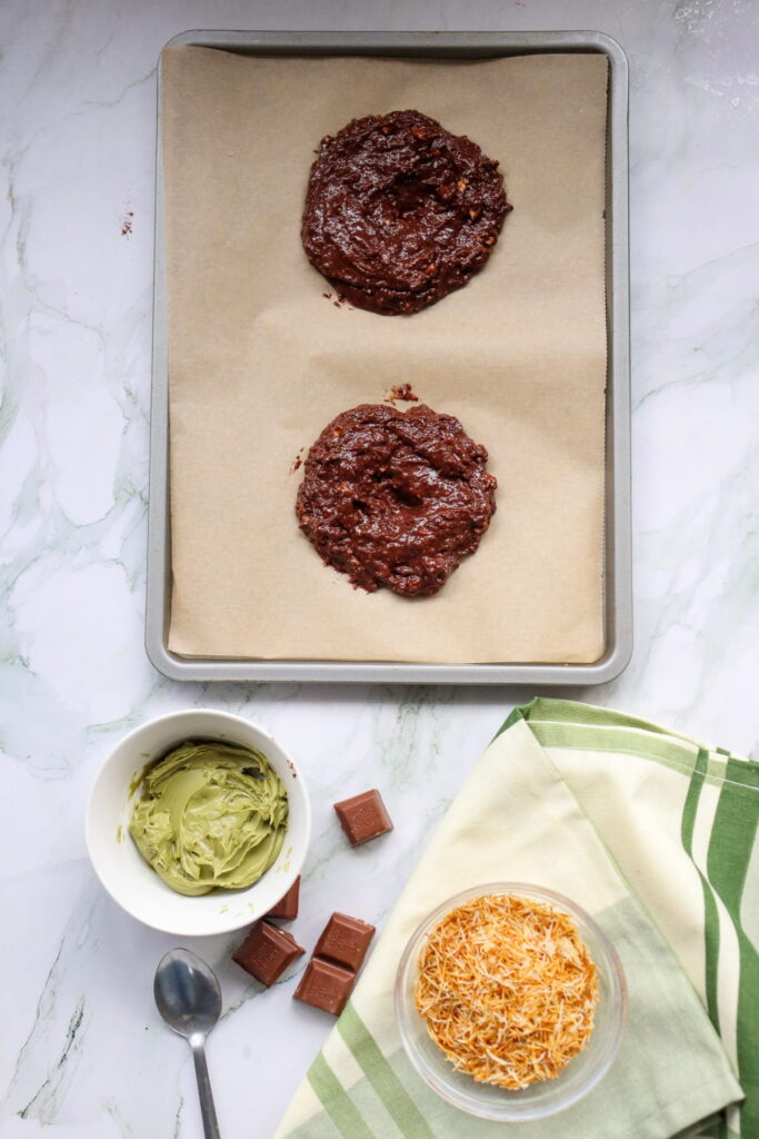 A baking sheet with two unbaked chocolate cookie dough portions on parchment paper; nearby are a bowl of green frosting, chocolate pieces, shredded cheese, and a spoon.