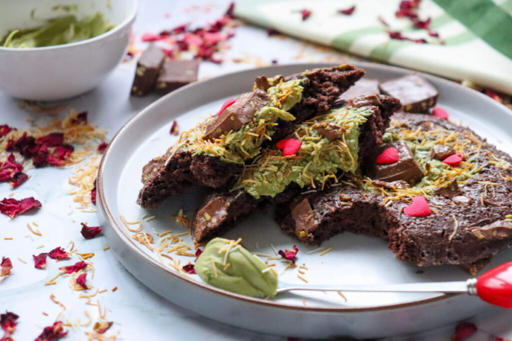 A plate with slices of chocolate dessert topped with green cream, chocolate pieces, red heart sprinkles, and scattered dried rose petals and grated garnish.