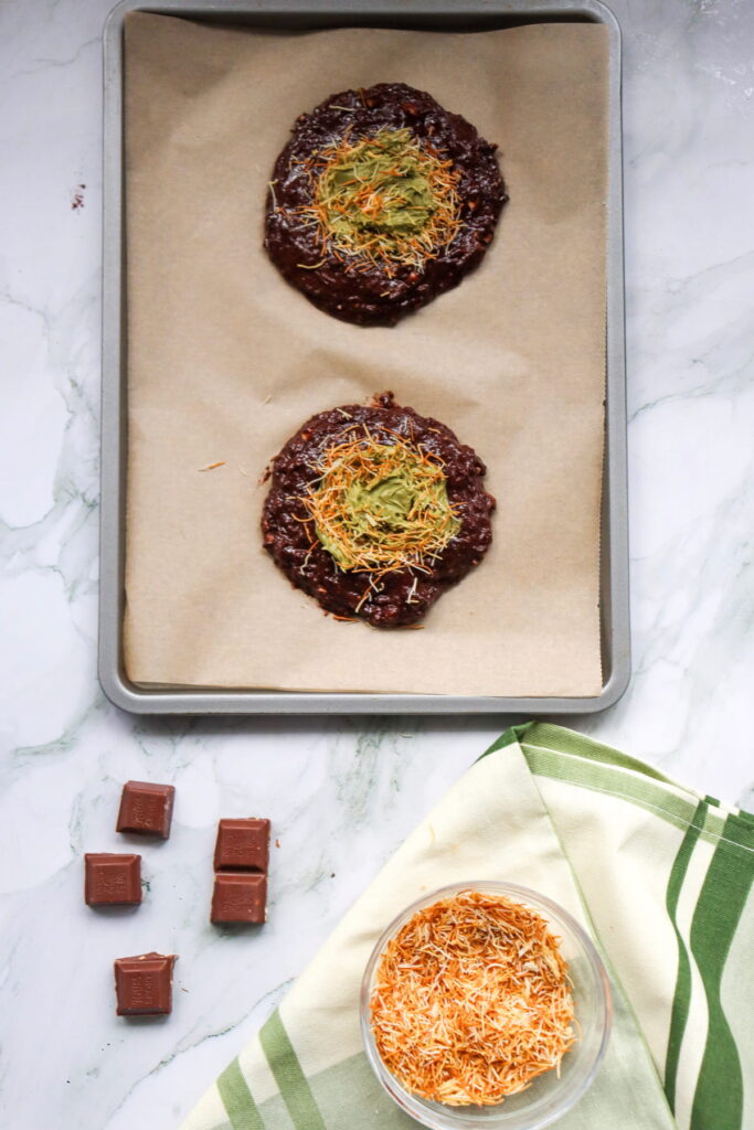 A baking sheet with two unbaked chocolate cookies topped with toasted shredded coconut and green filling, next to chocolate squares and a bowl of toasted coconut on a marble surface.