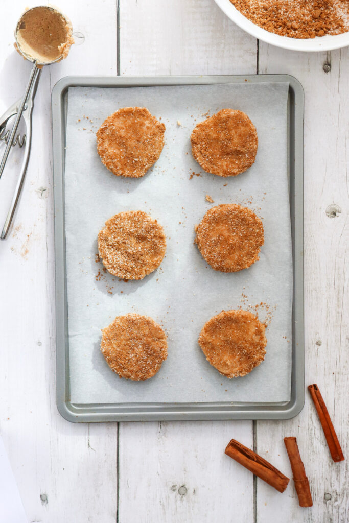 A baking sheet with six round, uncooked cookie dough portions coated in cinnamon sugar, set on parchment paper. A mixing bowl, scoop, and cinnamon sticks are nearby on a white wooden surface.