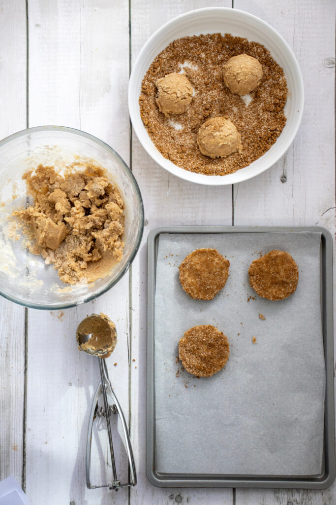 Cookie dough balls being rolled in sugar, with some on a baking sheet lined with parchment paper, and a bowl of dough and a scoop nearby on a white wooden surface.