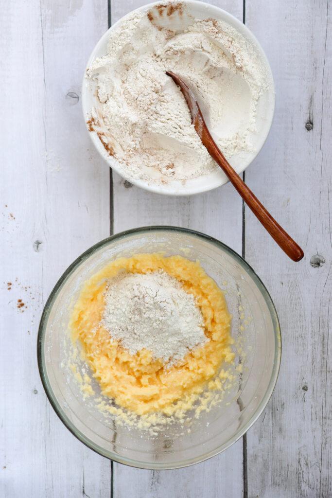 Two bowls on a wooden surface: one with flour and a wooden spoon, the other with creamed butter and sugar topped with added flour.