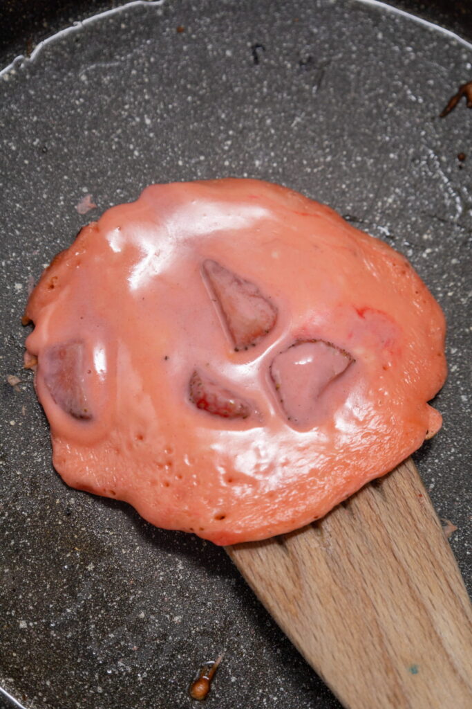A melted pink substance with chunks, possibly candy or fruit, sits on a wooden spatula in a frying pan.