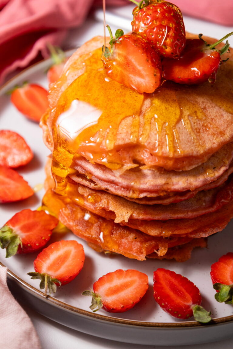 A stack of pancakes topped with strawberries, a pat of butter, and syrup, surrounded by sliced strawberries on a white plate.