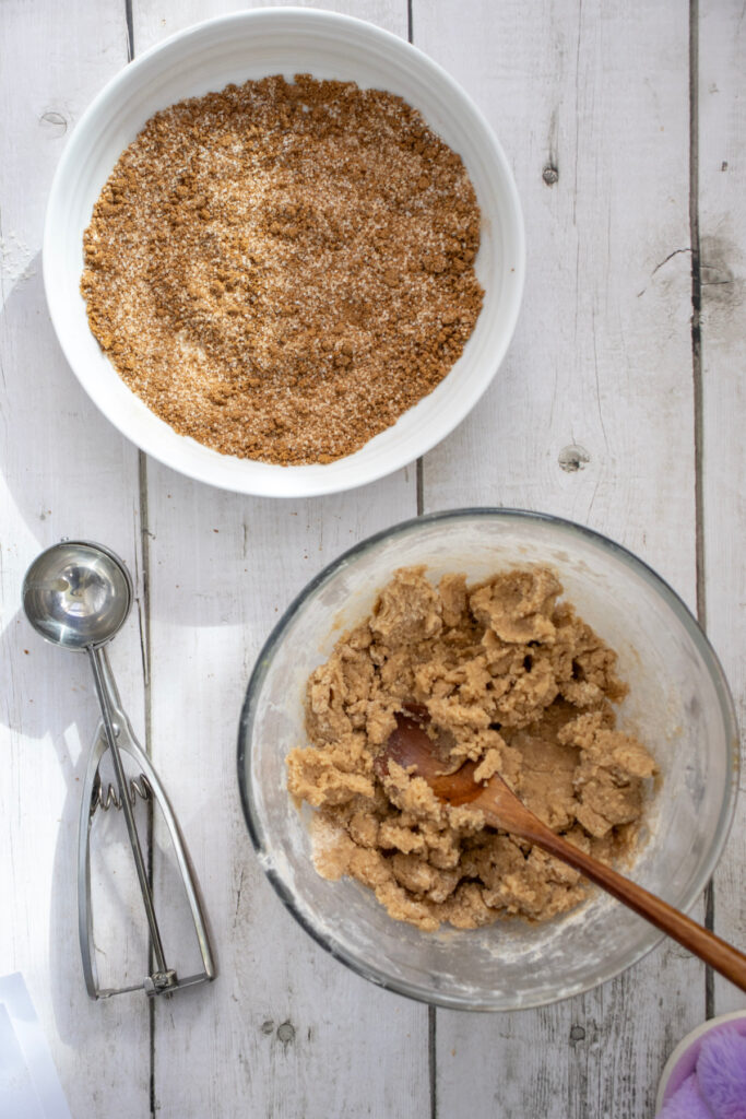 Two bowls on a white wooden surface: one with brown sugar and cinnamon mixture, the other with cookie dough and a wooden spoon. A metal cookie scoop lies beside the bowls.