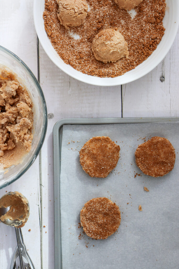 Cookie dough balls are rolled in cinnamon sugar, then placed on a parchment-lined baking sheet and flattened, ready for baking. Nearby are a mixing bowl and a dish with leftover dough and sugar.