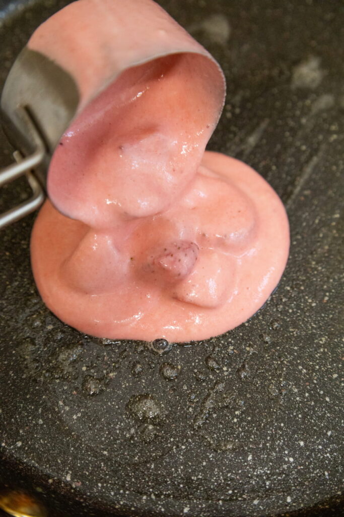 A close-up of pink batter being poured from a metal measuring cup onto a black nonstick pan.