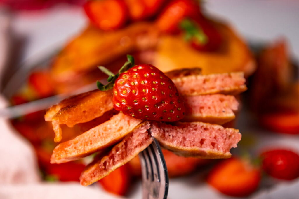 A fork holds pieces of pink pancakes topped with a fresh strawberry, with more pancakes and strawberries blurred in the background.