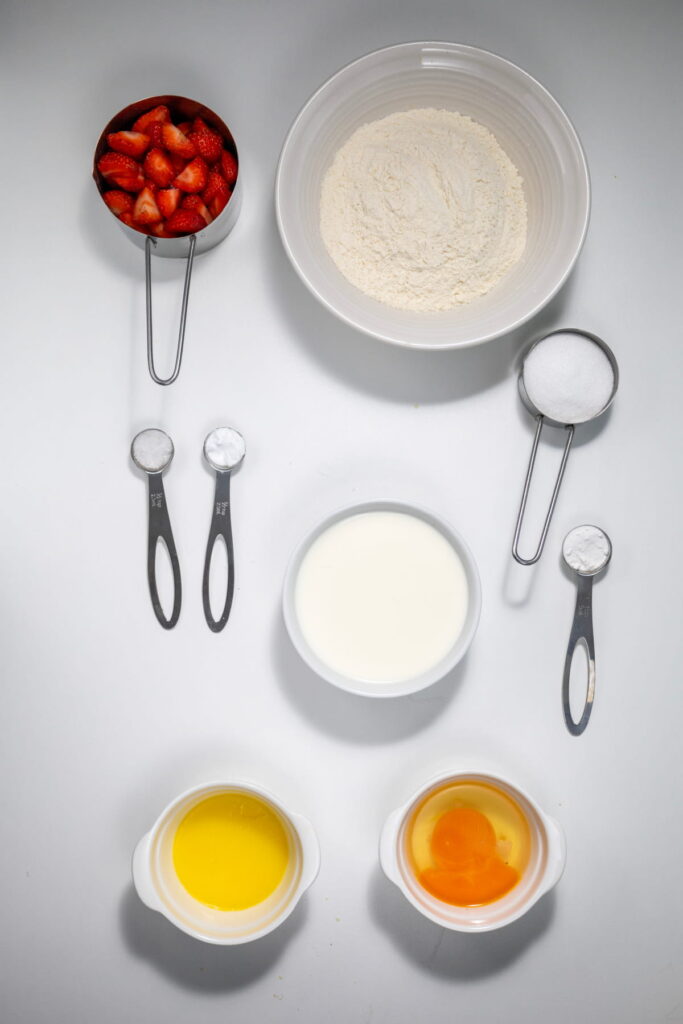 Overhead view of baking ingredients: sliced strawberries, flour, sugar, salt, baking powder, baking soda, milk, melted butter, and two eggs arranged on a white surface.
