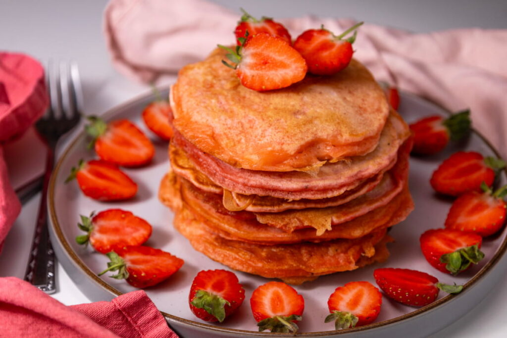 A stack of pink pancakes topped with sliced strawberries, surrounded by more strawberries on a plate. Pink and red serviettes are placed nearby.