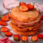 A stack of pink pancakes topped with sliced strawberries, surrounded by more strawberries on a plate. Pink and red serviettes are placed nearby.