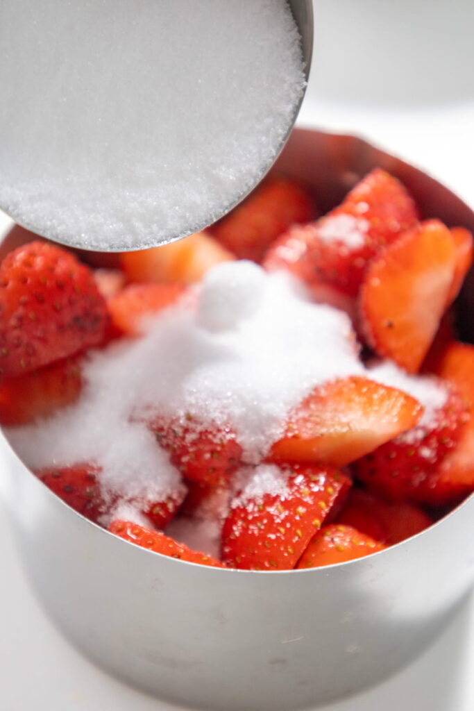 A tablespoon of granulated sugar is being poured over sliced strawberries in a metal bowl.