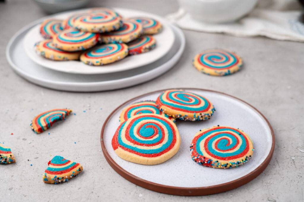 Colorful spiral cookies with red and blue swirls and sprinkled edges are arranged on two plates, with some broken pieces scattered on a light gray surface.