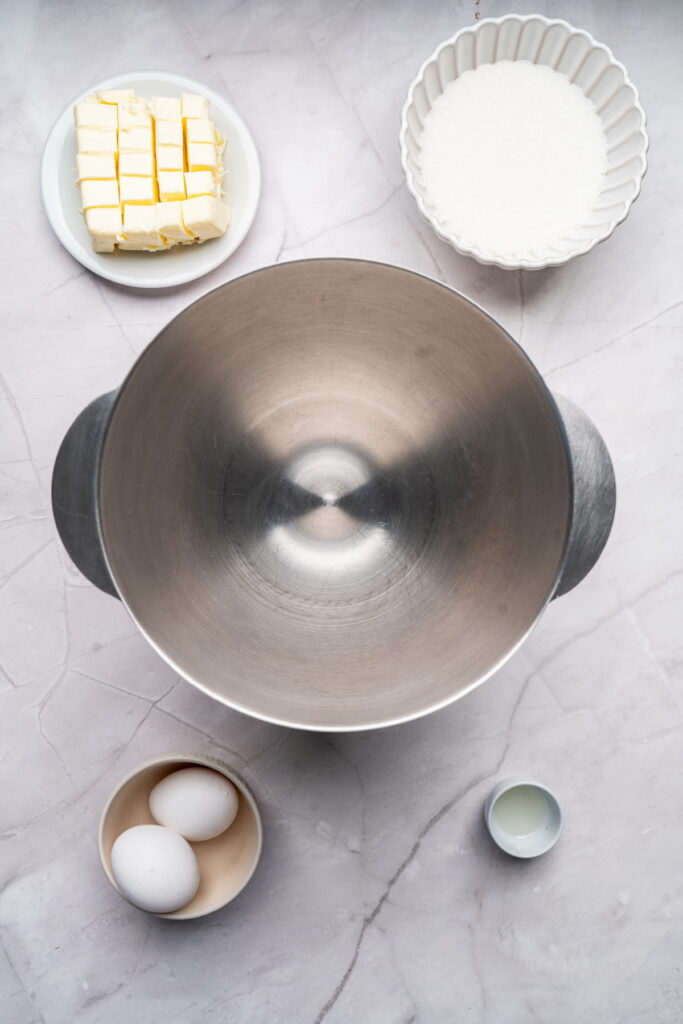 Overhead view of a metal mixing bowl surrounded by cubed butter, a bowl of sugar, two eggs, and a small bowl of liquid on a marble surface.