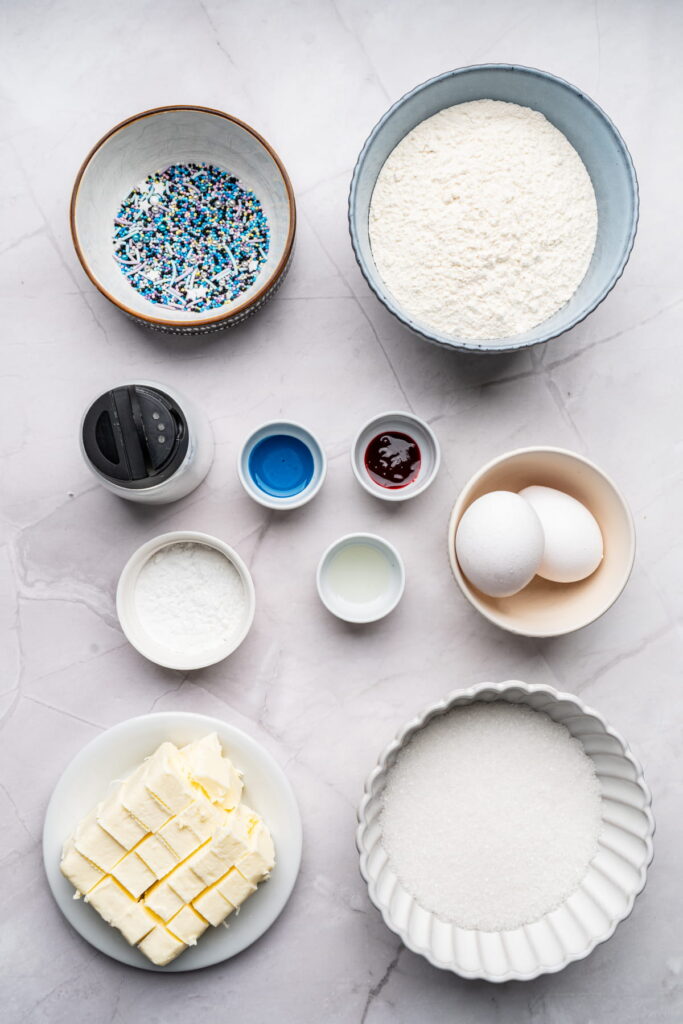 Assorted baking ingredients arranged in bowls and plates, including sprinkles, flour, sugar, butter, eggs, powdered sugar, food coloring, lemon juice, and vanilla extract.