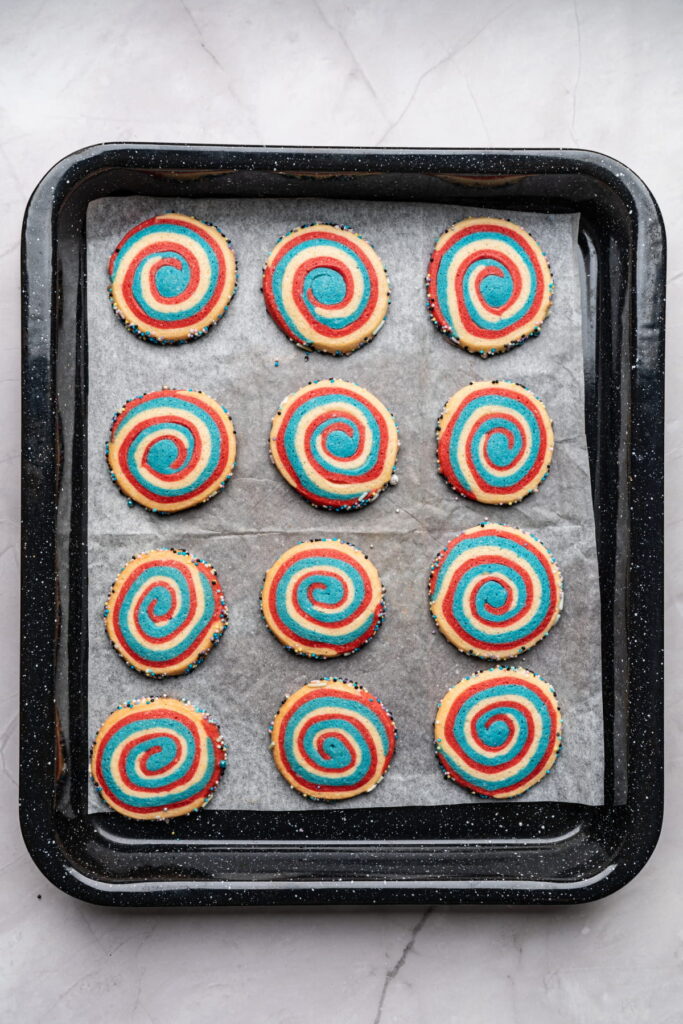 A baking tray with twelve colorful spiral cookies, featuring red, blue, and yellow swirls, arranged on parchment paper.