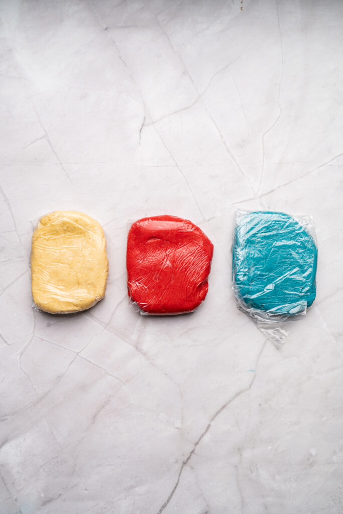 Three flattened discs of dough—yellow, red, and blue—wrapped in plastic wrap, placed side by side on a light marble surface.