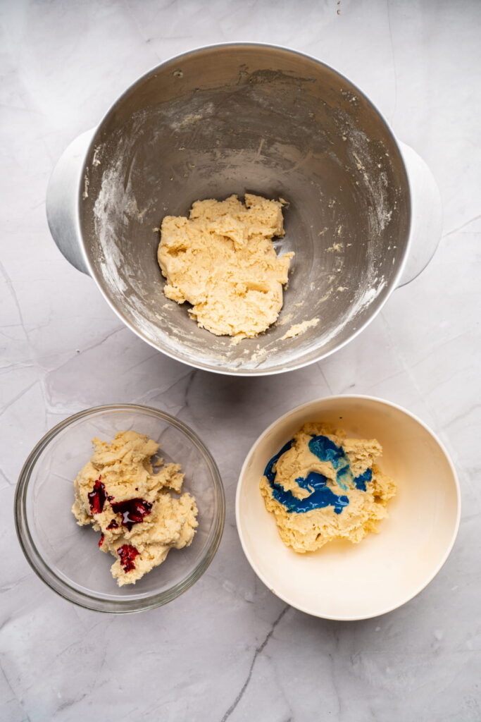Three bowls on a marble surface: one with plain cookie dough, one with dough and red food coloring, and one with dough and blue food coloring.