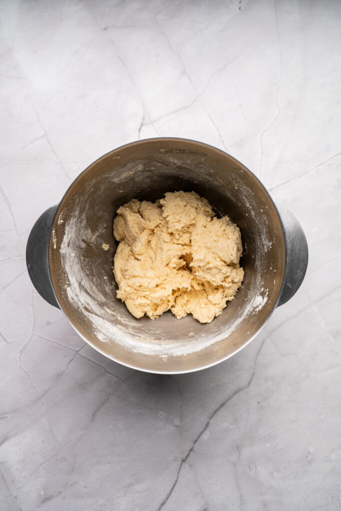 A metal mixing bowl containing a portion of raw dough sits on a light gray marble countertop.