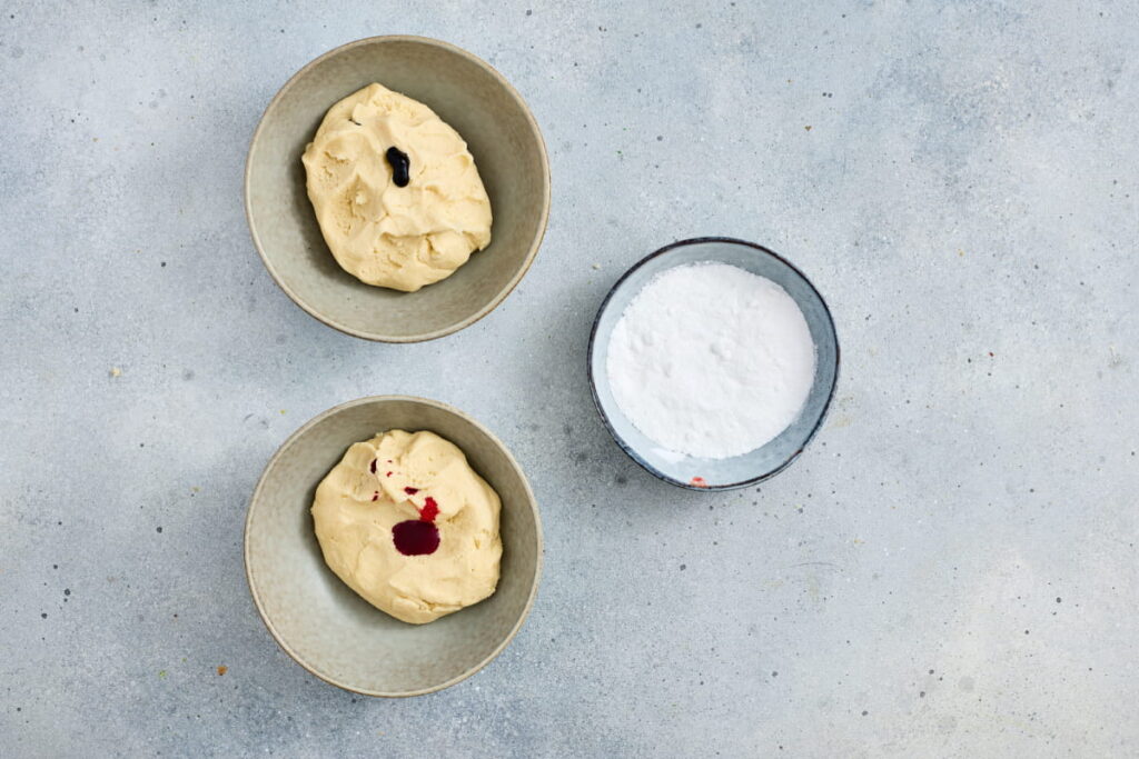 Two bowls of dough, each with food coloring on top, are next to a bowl of white powdered sugar on a light gray surface.