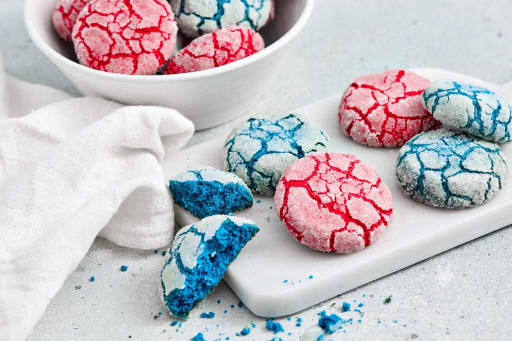 Red and blue crinkle cookies are displayed on a white surface, with some cookies on a small tray and others in a bowl, next to a white cloth.