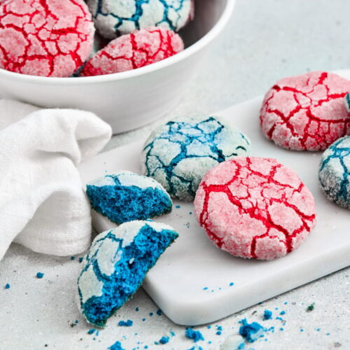 Red and blue crinkle cookies are displayed on a white surface, with some cookies on a small tray and others in a bowl, next to a white cloth.
