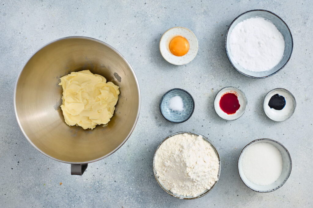 A mixing bowl with butter and small bowls containing an egg yolk, flour, sugar, powdered sugar, salt, and red and black food coloring on a gray surface.