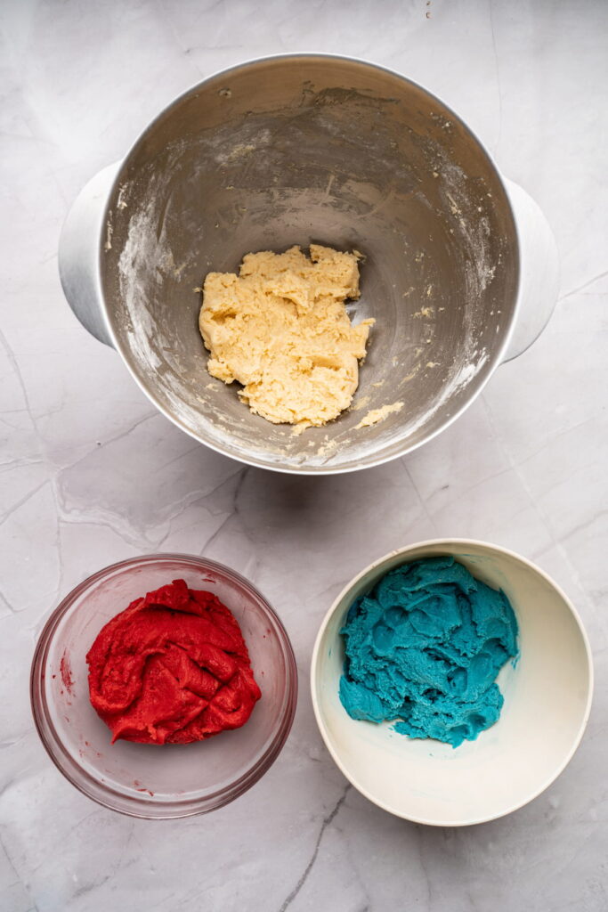 Three bowls of cookie dough on a marble surface: one with plain dough, one with red dough, and one with blue dough.